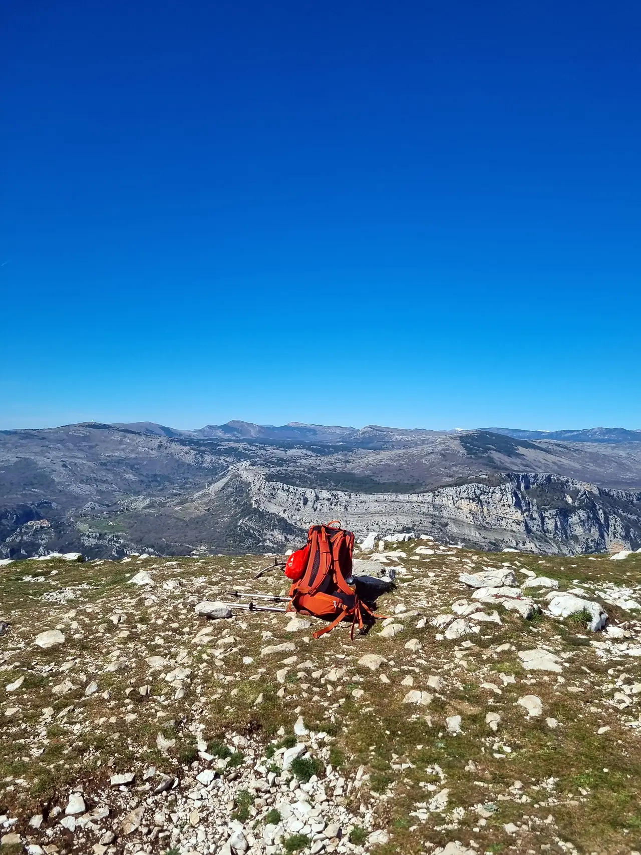 Un sac à dos orange posé au sommet, face à des montagnes calcaires sous un ciel bleu, photographié par Christel Louvet